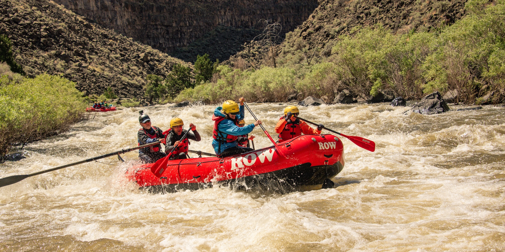 Group of rafters paddling through whitewater rapids in a scenic canyon in the USA, wearing helmets and life jackets.