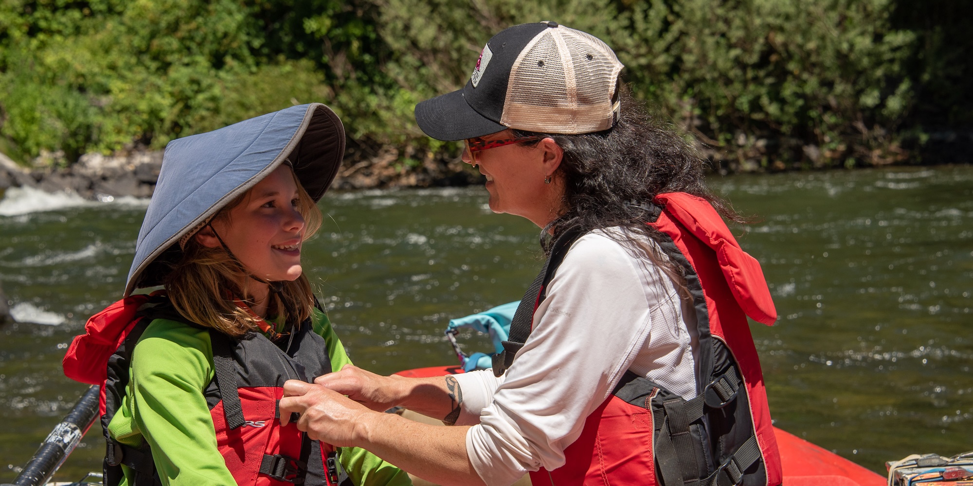 Guide helps a smiling child secure their life jacket before a rafting trip, both wearing sun hats and standing on a red raft.