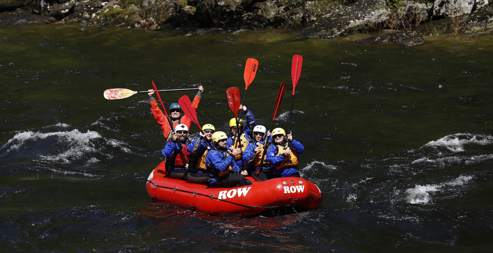 Happy group of rafters dressed in cold-weather gear holding up paddles in celebration on a calm stretch of river.