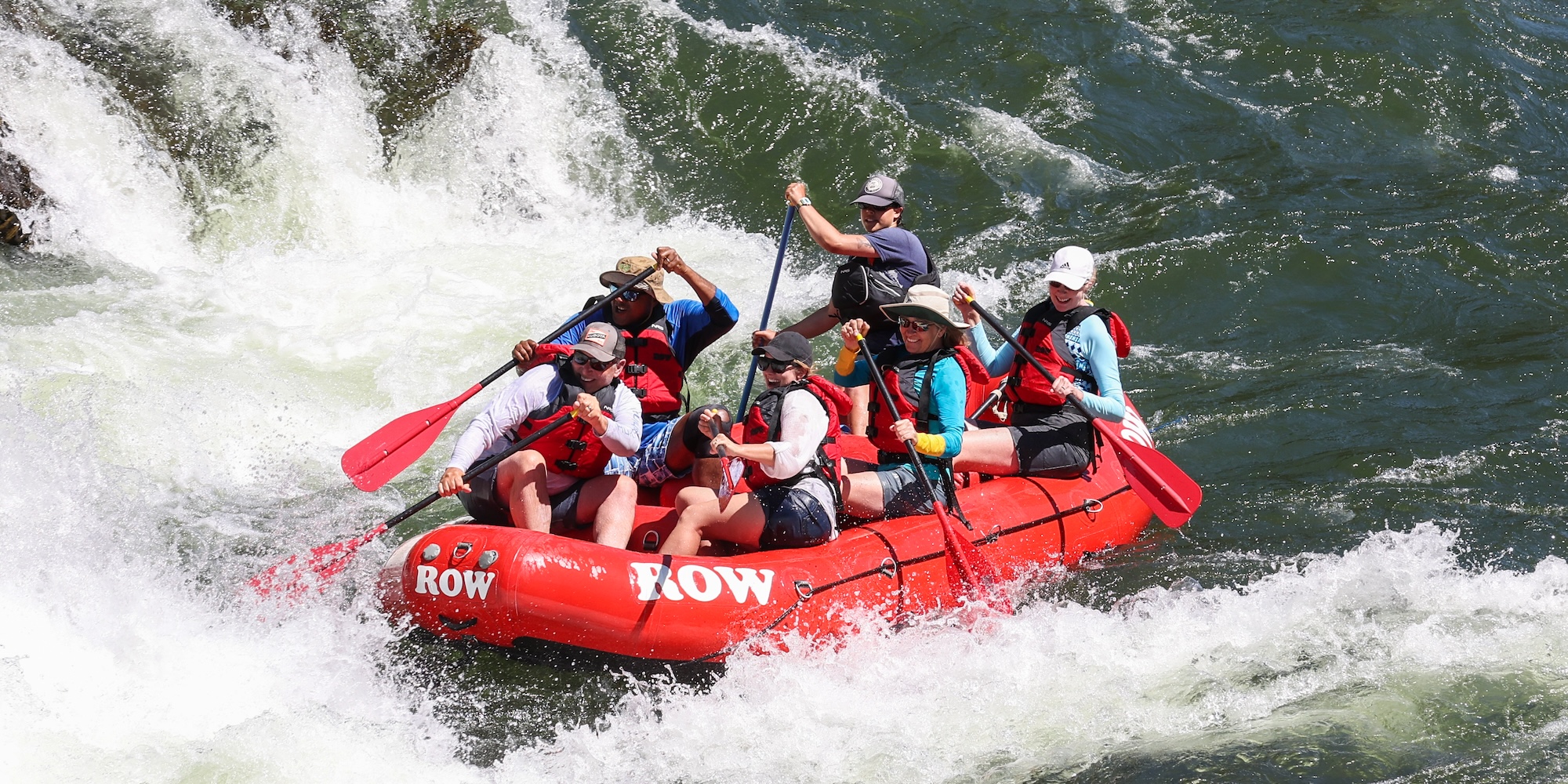 Excited group of paddlers tackling rapids in Montana, smiling and paddling together in a red raft.