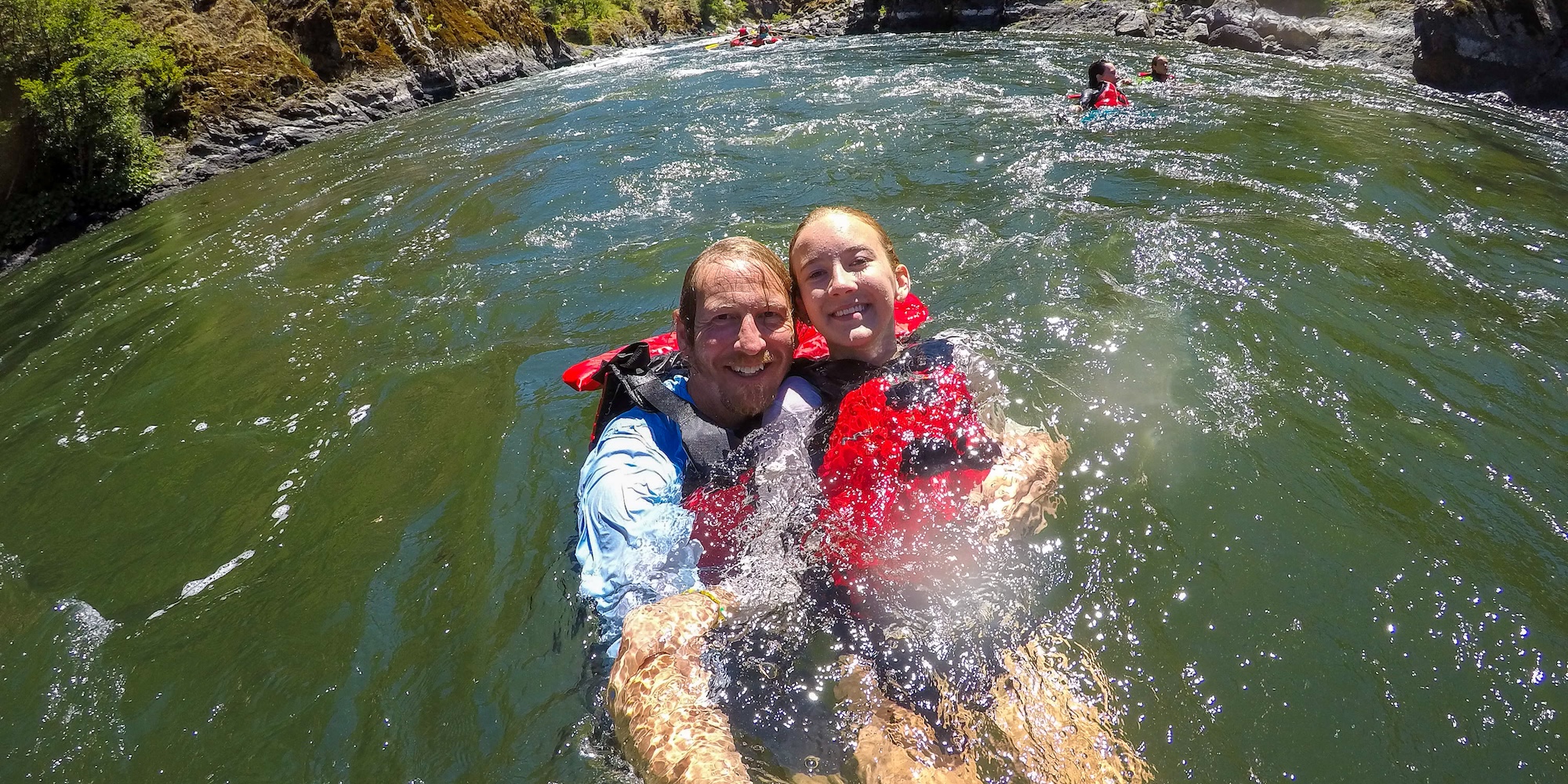 Father and daughter smiling while floating in a calm river with life jackets during a rafting trip swim break.