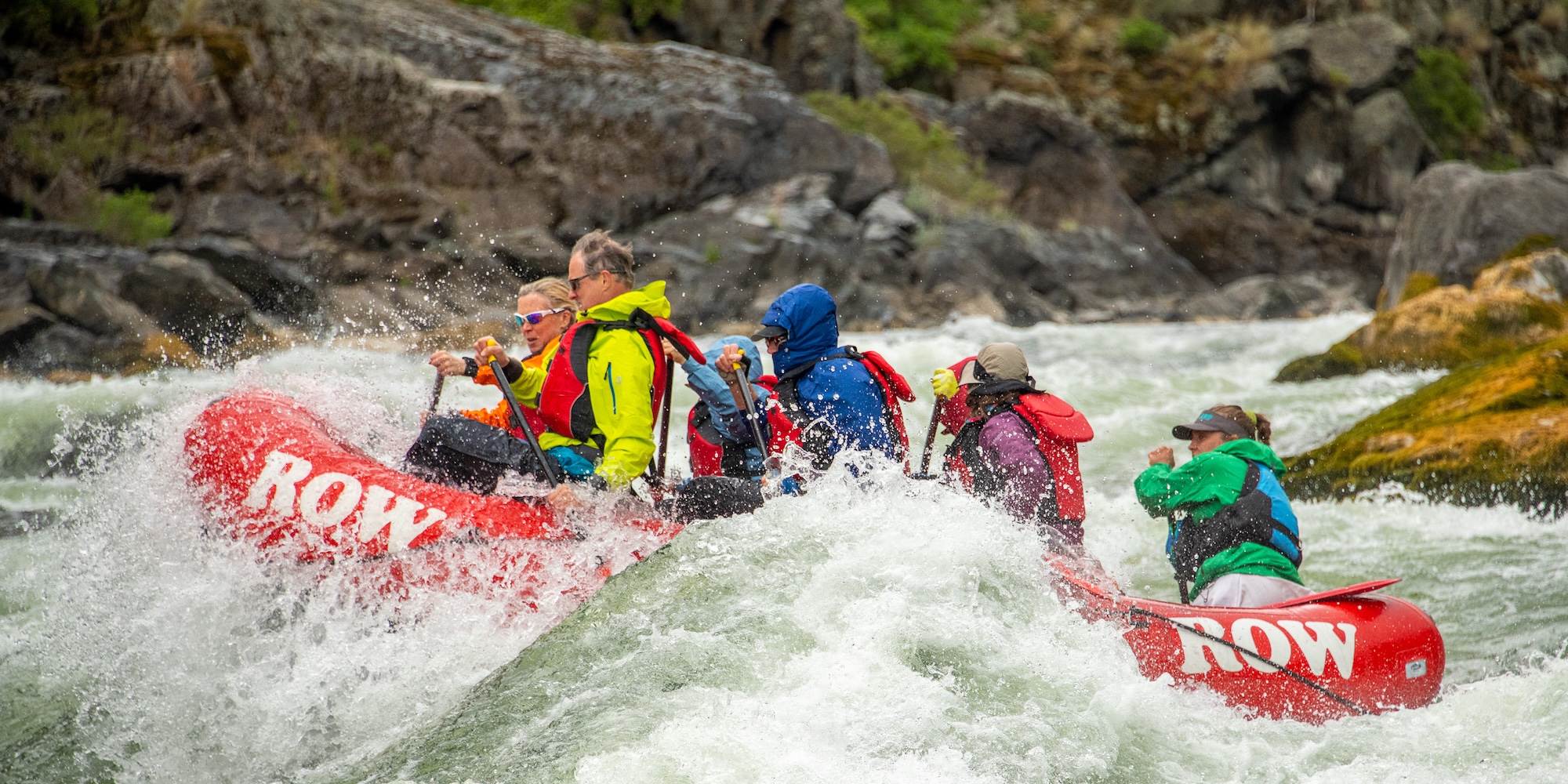 Group of rafters dressed in colorful cold-weather gear paddling through a powerful rapid in a red ROW Adventures raft, surrounded by rocky canyon walls.