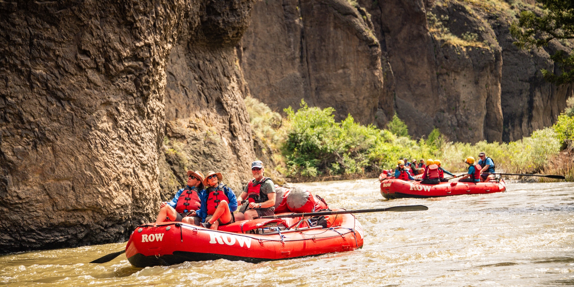 Rafters navigating calm water between canyon walls on a guided whitewater rafting trip, with multiple red rafts in view.