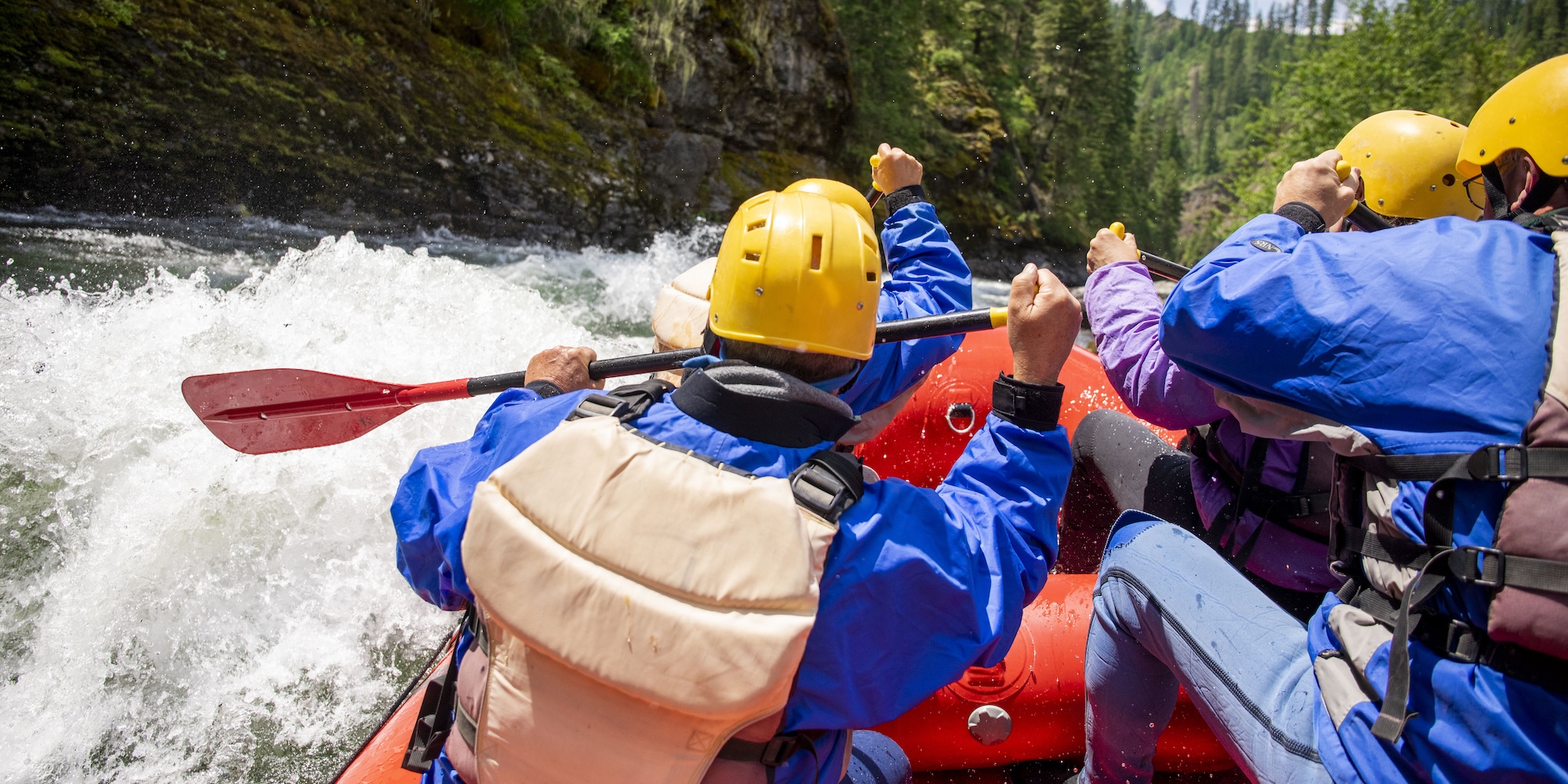 Close-up view of rafters in splash gear paddling through whitewater, water spraying over the side of the raft as they move forward.