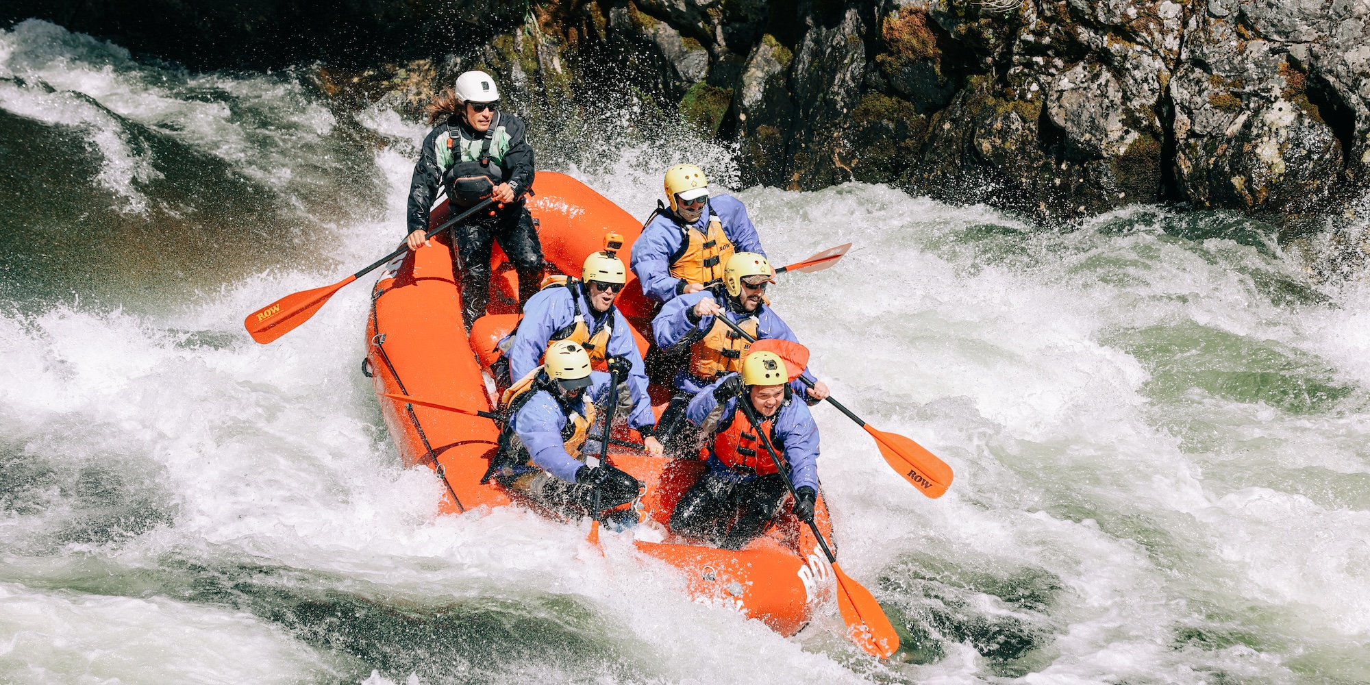 Group of rafters in blue jackets and yellow helmets navigating a powerful rapid on the Lochsa River in a bright orange raft.