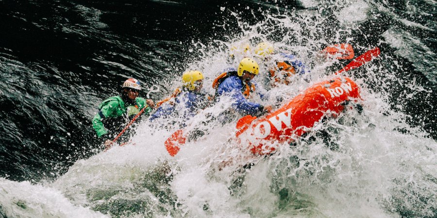 A red raft splashing through a large wave while all of the guests are sprayed with water. 