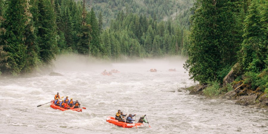 Two red rafts filled with guests floating down a misty river surrounded by dense, green forest. 
