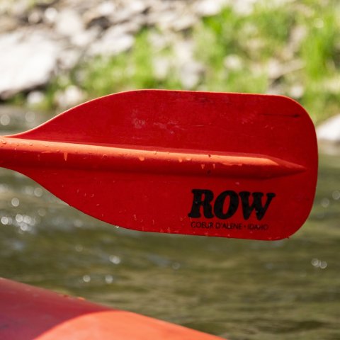 Close-up of a bright red ROW Adventures paddle resting on the side of a raft during a calm stretch of river.