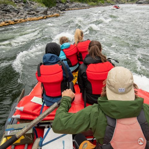 Four people and one river guide in a whitewater raft navigating downstream in a river.